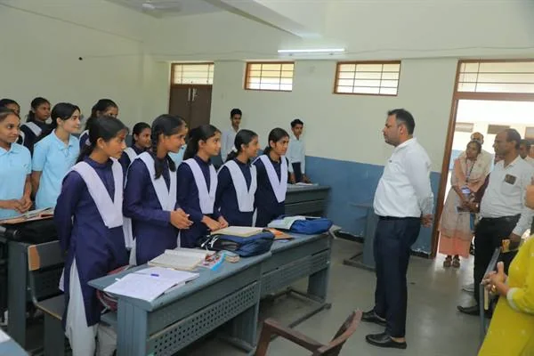 कक्षा में कतार से खड़ी छात्राओं से बातचीत करते कलेक्टर, सामने डेस्क पर किताबें और बैग रखे हैं