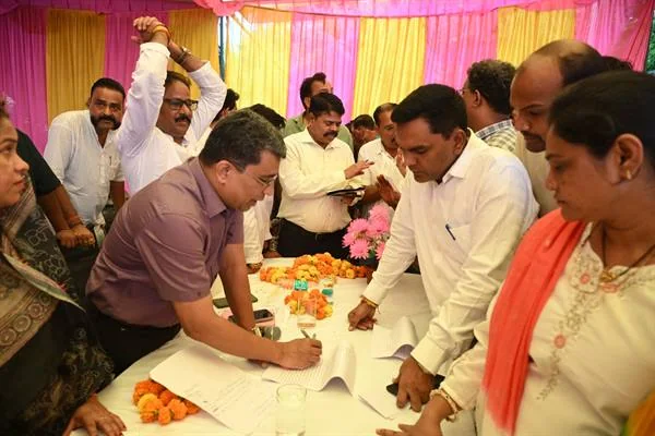 सजे हुए पंडाल में मेज पर रखे दस्तावेज पर लिखता एक व्यक्ति और आसपास खड़े लोग